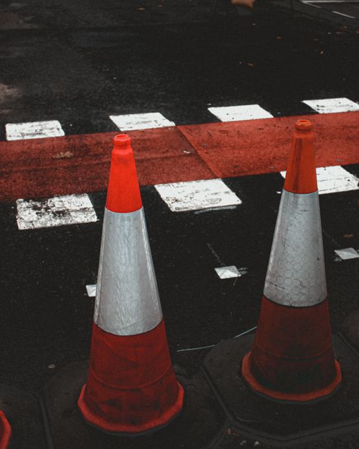 The image shows two orange and white traffic cones placed on a dark, asphalt road near a pedestrian crossing with white dashed lines and a solid red painted section. The cones appear weathered, with v