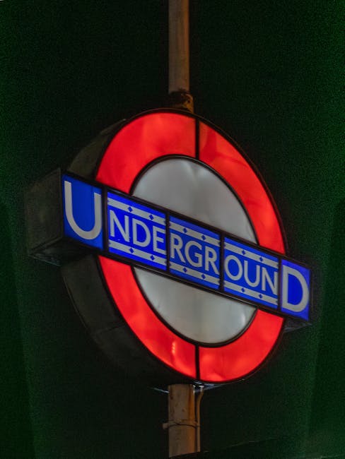 A close-up view of the iconic London Underground roundel sign illuminated at night, featuring a red circular border with a white centre and a blue rectangular bar displaying the word 'UNDERGROUND' in