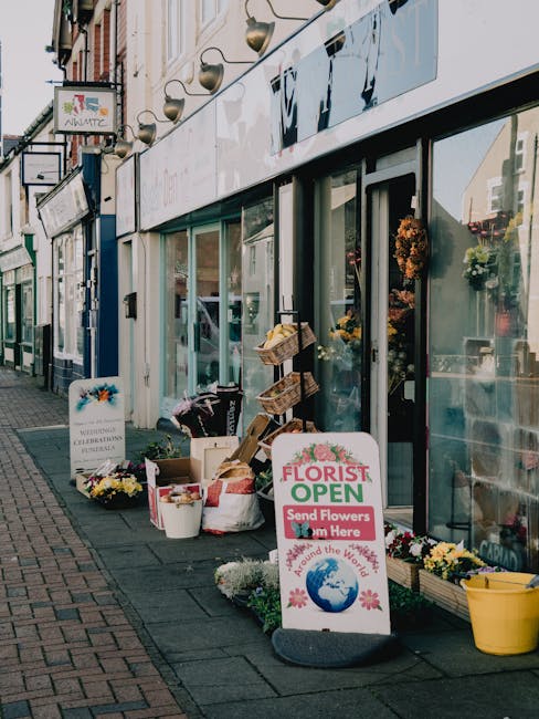 Outside Rayners Lane Florist, a flower shop with large glass windows and a white sign indicating the store is open, decorated with pink and green floral graphics. In front, an assortment of flower arr
