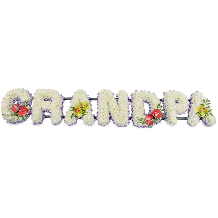 In this image, a bespoke funeral flower tribute spells the word "GRANDPA" in large, three-dimensional block capitals that run horizontally across a clean white backdrop. Each letter is meticulously covered in a dense mat of small white chrysanthemums, creating a rich, cushion-like effect where no stems or gaps are visible. The soft, rounded petals catch the light and combine to give the tribute a calm, even glow, suggesting purity, comfort, and peace. Framing the base of each letter is a narrow band of pale lavender ribbon, which just peeks out from under the blooms and adds a delicate, pastel border that gently emphasises each shape. Hidden behind the flowers, slender dark green bars link all the letters together, giving the impression of a single, continuous display that could be set on a stand at a funeral service in one of the churches or crematoria near Rayners Lane. To break the expanse of white and introduce points of emotion, the florist has inserted four accent clusters. On the left-hand 'G' and the central 'D', rich red anthuriums with their glossy, heart-shaped spathes and bold central spadices rise from polished green foliage, possibly accompanied by hints of tiny purple fillers, drawing the eye and symbolising deep love and remembrance. On the first and last 'A', soft sprays of yellow-green orchids unfurl their refined, exotic blooms among fresh leaves, adding a touch of sophistication and continuity. The tribute feels thoughtfully balanced and deeply personal, the kind of arrangement a family around Rayners Lane might choose to honour a grandfather who meant the world to them, offering a final message of respect and affection.